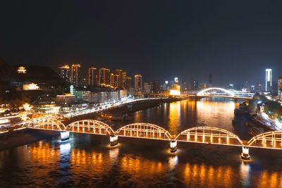 Bridge over river by illuminated buildings against sky at night