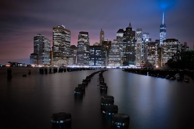 Wooden posts amidst river against cityscape at night