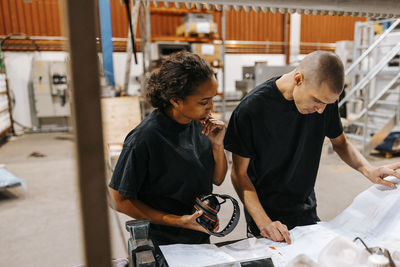 Mature male worker reading blueprint while standing next to female colleague holding wireless headphones in warehouse
