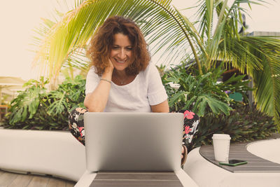 Smiling woman using laptop while sitting by plants outdoors