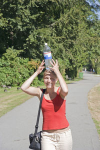 Portrait of young woman balancing water bottle on head at road during sunny day