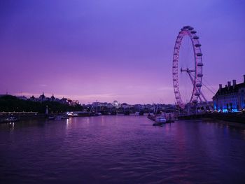 Illuminated ferris wheel by river against sky at dusk