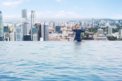 Happy boy with arms outstretched sitting on infinity pool against cityscape