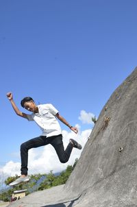 Low angle view of man jumping on rock against blue sky