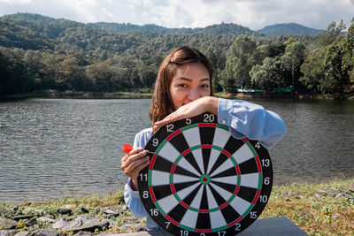 Portrait of young woman holding dart board by lake against sky