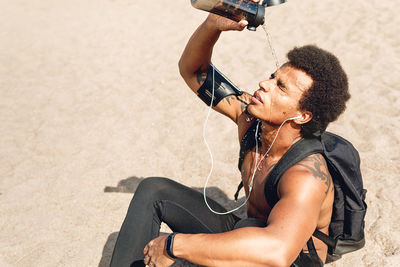 Shirtless athlete pouring water on face while sitting at beach