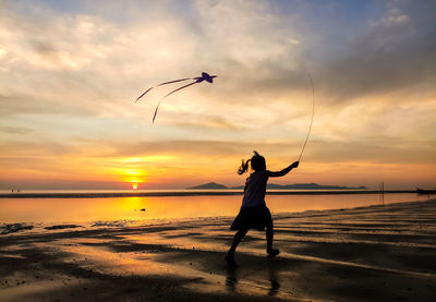 Silhouette girl flying kite at beach against sky during sunset