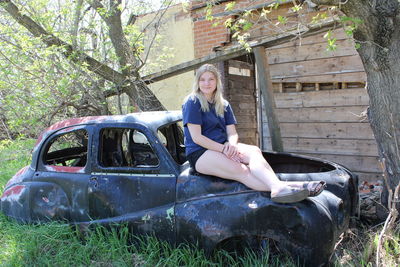 Portrait of smiling young woman sitting on wrecked vintage car