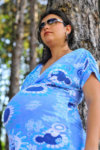 Pregnant woman wearing sunglasses looking away while leaning on tree