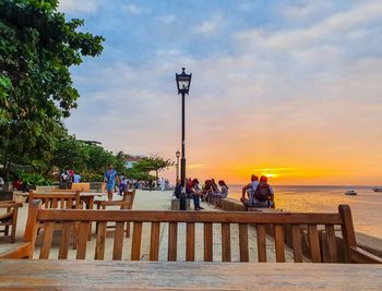 People sitting on street by sea against sky during sunset