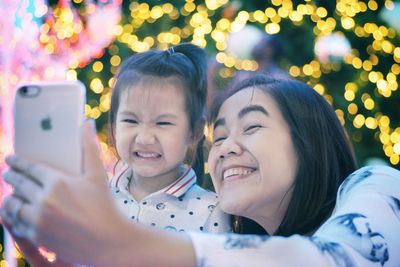 Smiling mother and daughter taking selfie with smart phone against illuminated christmas tree