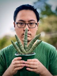 Portrait of young man holding fan shaped succulent plant in a pot against trees and cloudy sky.