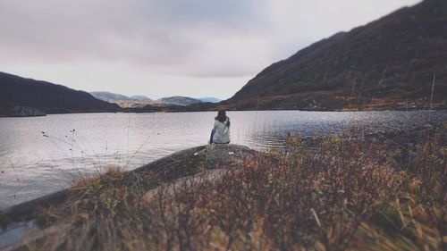 Rear view of woman sitting at lakeshore against cloudy sky