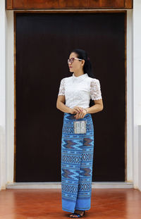 Full length of woman in traditional clothing holding lunch box while standing on tiled floor against wooden door