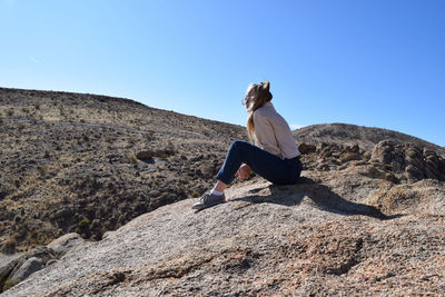 Woman sitting on rock against mountains against clear blue sky