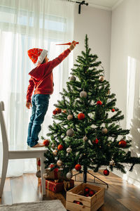 Boy in a santa claus hat decorates the new year tree at home, puts christmas toy on  top of the tree