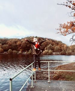 Man standing on railing by lake against sky