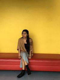 Young woman looking away while sitting on sofa against yellow wall