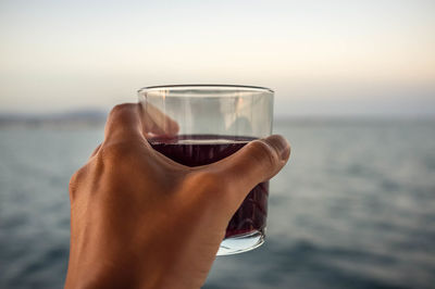 Close-up of hand holding drink against sea at sunset