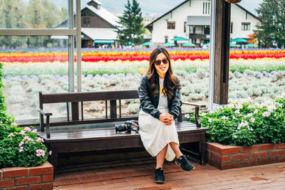 Full length portrait of young woman sticking out tongue while sitting on bench against field