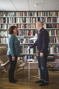 Full length of male therapist gesturing while talking with patient by bookshelf at home office