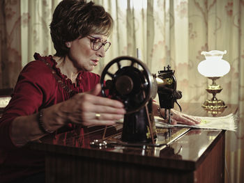 Senior lady in glasses using retro sewing machine to create linen napkin in cozy room at home