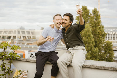 Carefree gay couple toasting drinks while sitting on wall during party in balcony