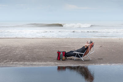 Mature man with gray hair resting with arms up on a beach chair. the sea in the background.