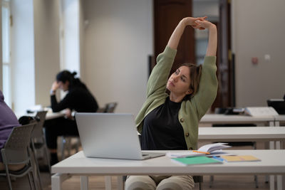 Relaxed middle-aged female stretching hands taking break from work on laptop in library or classroom