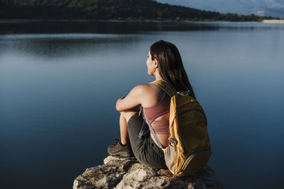 Young female backpacker with backpack sitting on rock by lake