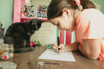 Cat watching girl drawing picture on paper at home