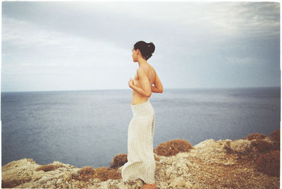 Side view of shirtless woman standing on rock formation against sky