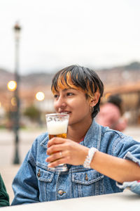 Delighted young woman sitting at table in veranda of pub in city of cusco and drinking glass of beer and looking away