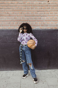 Female teenager with basketball standing against wall on footpath