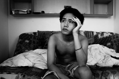 Thoughtful shirtless young man looking away while sitting on bed at home