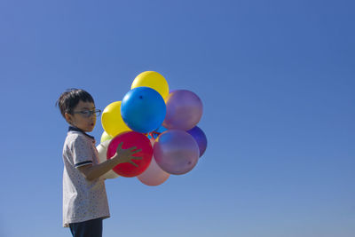 Boy holding multi colored balloons while standing against clear blue sky