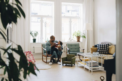 Full length of father feeding milk to daughter while sitting in living room at home