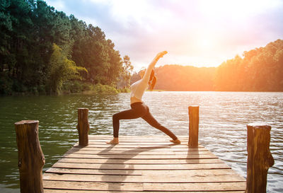 Woman doing yoga on pier over lake against sky during sunset