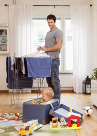 Father drying clothes while looking at baby boy playing with toys
