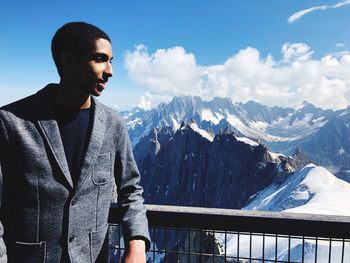 Young man standing by railing against snowcapped mountains and sky