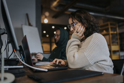 Young female hacker working on laptop by male colleague at desk in startup company