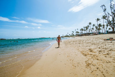 Rear view of shirtless man walking at beach against sky during sunny day