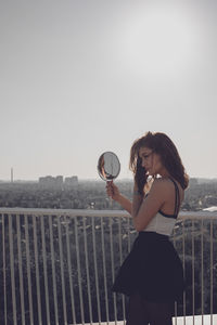 Woman holding mirror in city against clear sky