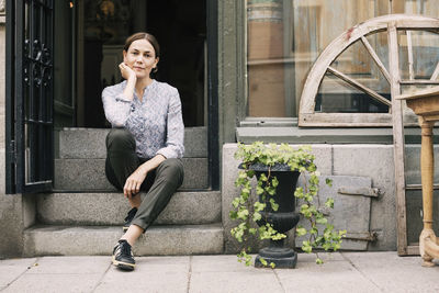 Portrait of female owner with hand on chin sitting on steps at store