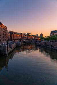 Bridge over river by buildings against sky during sunset