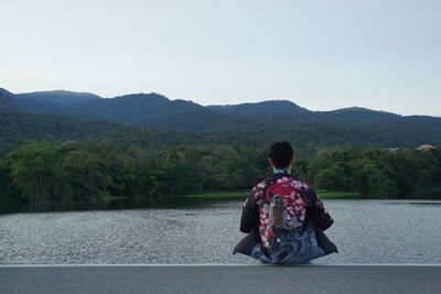 Rear view of man sitting by lake and mountains against sky