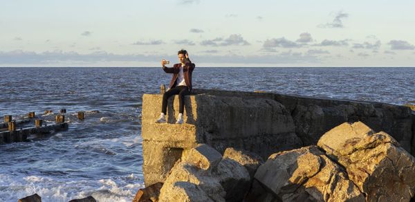 Man taking selfie while sitting on rock against sea and sky