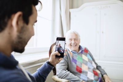 Caretaker photographing happy senior man through smart phone at nursing home