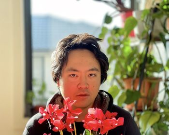 Close-up of young asian man holding red flowering cyclamen plant against house plants and window.