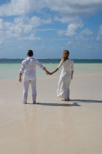 Rear view of couple holding hands while standing at beach against sky